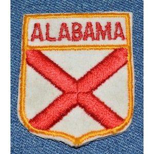 May include: A white and red embroidered patch with a yellow border. The patch features the word "ALABAMA" in red lettering and a red cross on a white background.