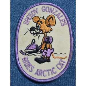 May include: A white oval embroidered patch with a purple border. The patch features a cartoon mouse wearing purple pants and a number "1" on its chest. The mouse is riding a purple snowmobile. The text "Speedy Gonzales Rides Arctic Cat" is embroidered around the edge of the patch.