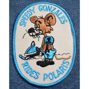 May include: A white oval embroidered patch with a blue border. The patch features a cartoon mouse wearing blue pants and a brown racing bib with the number "1" on it. The mouse is riding a blue snowmobile. The text "Speedy Gonzales Rides Polaris" is written around the edge of the patch.