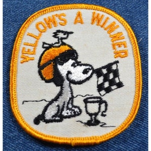 May include: Yellow embroidered patch with a cartoon dog wearing a helmet and holding a checkered flag. The text "Yellow's A Winner" is around the edge of the patch.