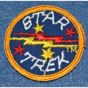 May include: A blue embroidered patch with a white outline. The patch features a yellow lightning bolt with red accents, five stars, and the text "STAR TREK" in white.