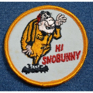 May include: A white embroidered patch with a yellow border. The patch features a cartoon character wearing a yellow jumpsuit and the text "Hi Snobunny".
