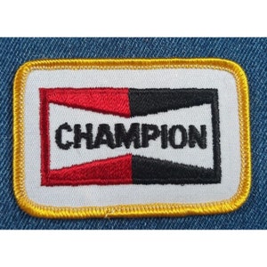 May include: A white embroidered patch with a red and black logo that says "CHAMPION" in black lettering. The patch has a yellow border.