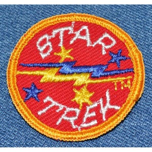 May include: A round, embroidered patch with the words "STAR TREK" in white, set against a red background. The design includes yellow and blue lightning bolts and stars, with an orange border. The patch is on a blue denim background.