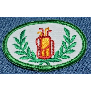 May include: A white oval embroidered patch with a green border. The center of the patch features a red and yellow golf bag with golf clubs sticking out of the top. The golf bag is surrounded by two green leafy branches.