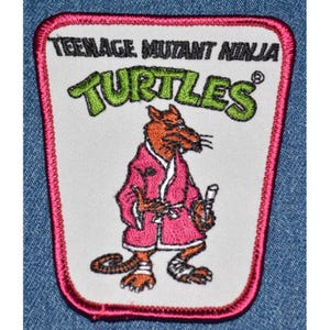 May include: Embroidered patch featuring the words "TEENAGE MUTANT NINJA TURTLES" in green and a cartoon rat wearing a pink robe. The patch has a white background with a pink border and is likely for clothing or accessories.