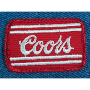 May include: Embroidered red patch with the word "Coors" in white script. The patch has two white horizontal stripes and a red border. The patch is rectangular and is set against a blue denim background.