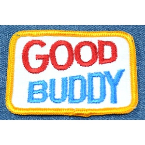 May include: A white embroidered patch with a yellow border. The words "GOOD BUDDY" are stitched in red and blue thread.
