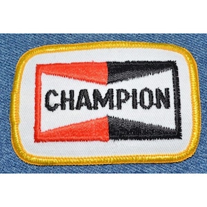 May include: A white embroidered patch with a red and black design. The patch has a yellow border and the word "CHAMPION" in black lettering.