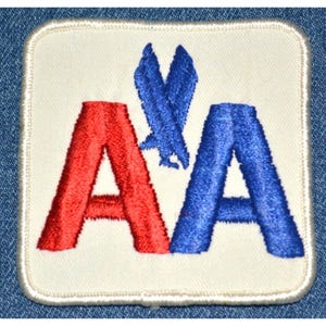 May include: A square patch with a white background features the letters "AA" in red and blue, with a blue stylized bird emblem between them. The patch has a white border and is likely an emblem or logo.