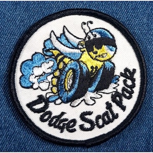 May include: A white, round embroidered patch with a black border. The patch features a cartoon bee with yellow and black stripes, blue wings, and a black eye mask. The bee is driving a car with blue wheels and white smoke coming out of the exhaust. The text "Dodge Scat Pack" is written in black below the bee.