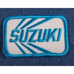 May include: A white embroidered patch with a blue outline and the word "SUZUKI" in blue lettering. The background of the patch has a blue sunburst design.