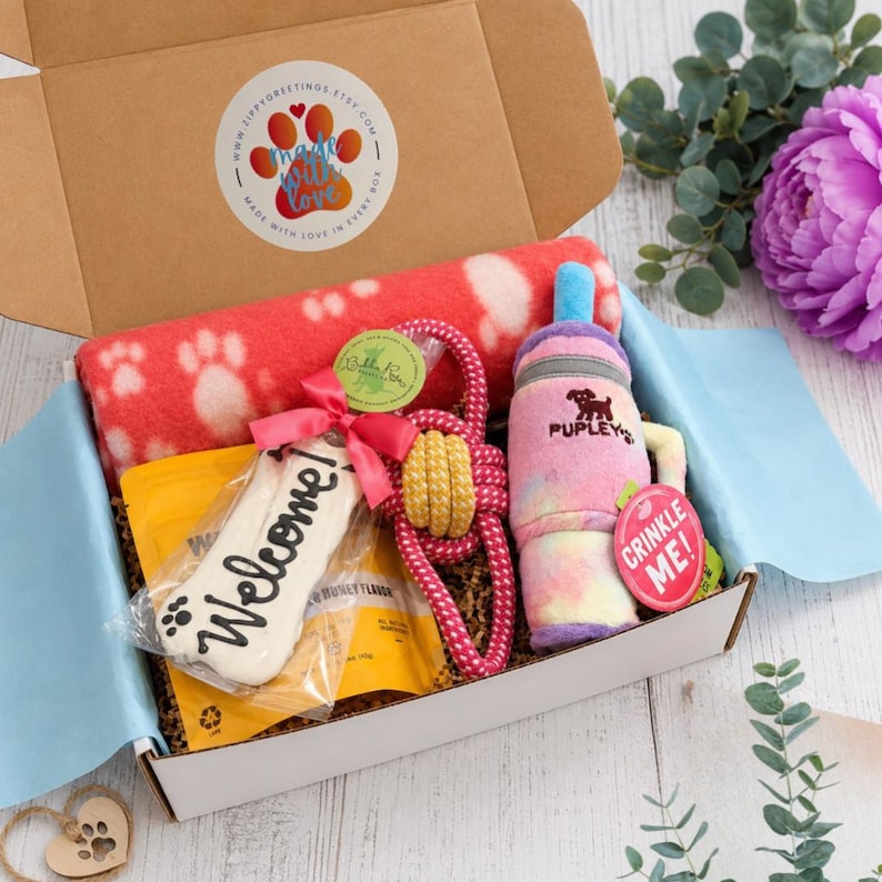 May include: A cardboard gift box containing a red and white paw print fleece blanket, a bone-shaped cookie labeled Welcome, a pink and yellow rope toy, and a plush bottle toy labeled Pupley with a pink tag that reads Crinkle Me.