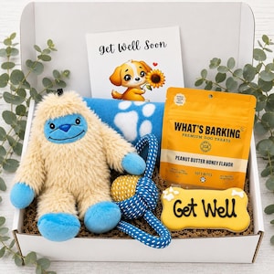 May include: A gift box containing a beige and blue plush yeti toy, a blue blanket with white paw prints, a yellow bag of What's Barking peanut butter honey dog treats, a bone-shaped cookie labeled Get Well, and a card with a puppy illustration.