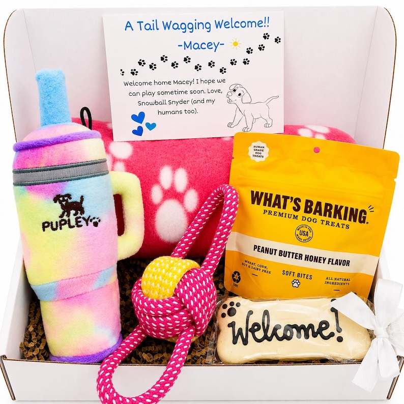 May include: A gift box containing a tie-dye plush tumbler toy labeled Pupley, a pink fleece blanket with white paw prints, a yellow and pink rope toy, a bag of What's Barking peanut butter honey dog treats, and a bone-shaped cookie labeled Welcome.