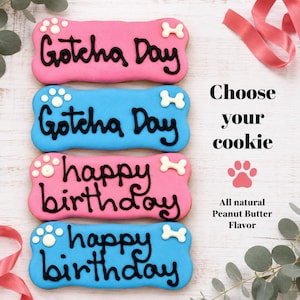 May include: Four bone-shaped dog cookies with pink or blue icing and black lettering. Two cookies read Gotcha Day and two read happy birthday. Text on the right reads Choose your cookie and All natural Peanut Butter Flavor with a paw print.