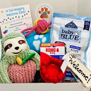 May include: A gift box for a puppy containing a plush sloth toy, a pink and yellow rope toy, a blue blanket with white paw prints, a bag of Blue Buffalo Baby Blue soft biscuits, a red Kong Ring, and a bone-shaped cookie with the word Welcome.