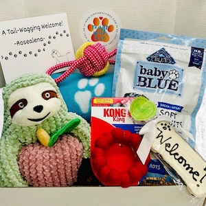 May include: A gift box for a dog, containing a plush sloth toy, a red Kong toy, dog treats, a rope toy, and a welcome bone-shaped treat. The box includes a card that says "A Tail-Wagging Welcome!"