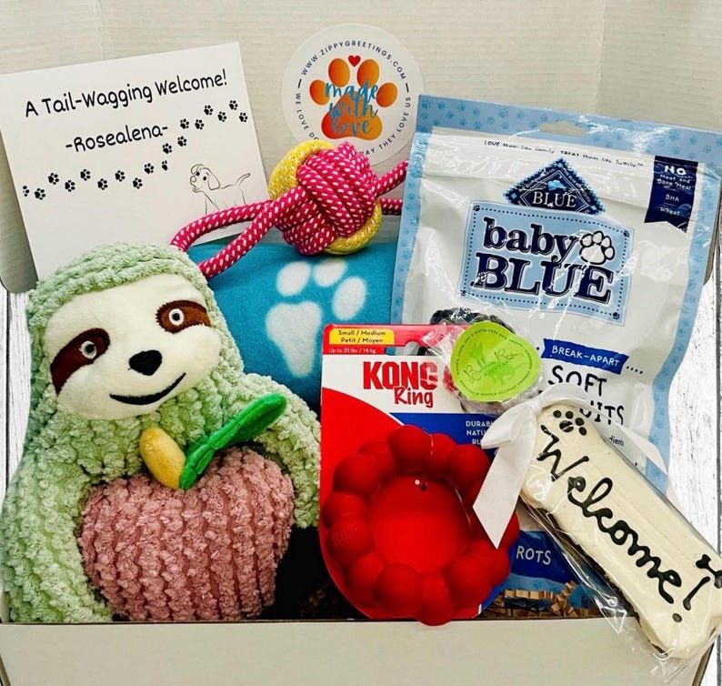 May include: A gift box for a dog, containing a plush sloth toy, a red Kong toy, dog treats, a rope toy, and a welcome bone-shaped treat. The box includes a card that says "A Tail-Wagging Welcome!"