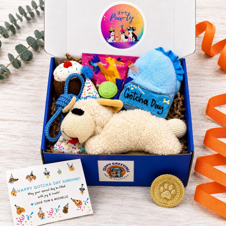May include: A blue gift box filled with dog toys including a plush cream-colored dog, a blue plush toy with the text Gotcha Day, a blue rope toy, a cupcake toy, and a party hat. A greeting card reads Happy Gotcha Day Sunshine.