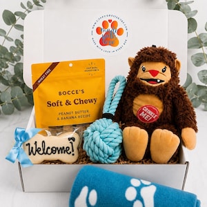 May include: A white gift box containing a yellow bag of Bocce's Soft & Chewy peanut butter and banana dog treats, a light blue rope toy, a brown plush monster toy with a red Crinkle Me tag, a bone-shaped cookie with Welcome written on it, and a blue blanket.