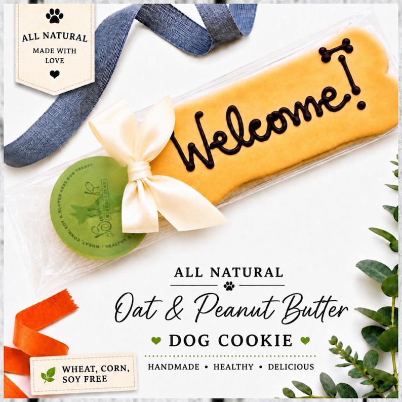 May include: A bone-shaped dog cookie in a clear plastic sleeve with a cream-colored ribbon. The cookie features the word Welcome in dark icing. A green circular label is attached, and text on the image reads All Natural Oat & Peanut Butter Dog Cookie.