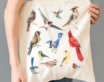 May include: Cream-colored tote bag featuring watercolor illustrations of various birds, including a red cardinal, blue jay, and hummingbird. The birds are depicted in flight and perched, showcasing a variety of colors and patterns. The bag has two handles and is held by a person.