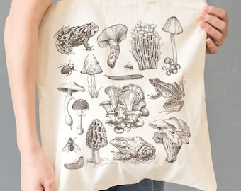 Puede incluir: Una bolsa de lona blanca con una ilustración en blanco y negro de varios hongos, ranas e insectos. La ilustración incluye una variedad de tipos de hongos, como rebozuelos, colmenillas y setas shiitake.