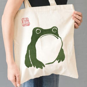 May include: Cream-coloured canvas tote bag with a large green frog design. The frog has a wide open mouth and large eyes. A red stamp is in the upper left corner. The bag is held by a person.