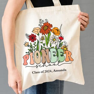Peut inclure: Un sac fourre-tout en toile blanche avec un motif floral et le texte "Pioneer School" en couleurs arc-en-ciel. Le texte "Class of 2024, Amanda" est imprimé sous le nom de l'école.
