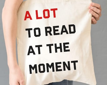 Puede incluir: Bolso tote de color crema con el texto "A LOT TO READ AT THE MOMENT" en letras negras y rojas. El bolso es sostenido por una persona. El bolso tiene un diseño sencillo y probablemente está hecho de lona o un material similar.