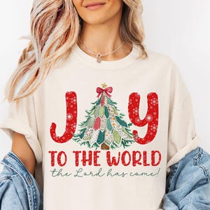 May include: Ivory-colored t-shirt with the words "Joy to the World" in red, with a Christmas tree design. The text "the Lord has come!" is below the main text. The shirt is part of the Oak & Olive Boutique collection.