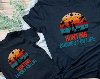 May include: Two black t-shirts with a vintage sunset graphic and the text "Hunting Buddies for Life". The larger shirt has a long sleeve and the smaller shirt has a short sleeve.