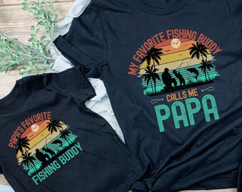 May include: Two black t-shirts with a retro sunset graphic and the text "Papa's Favorite Fishing Buddy" and "My Favorite Fishing Buddy Calls Me Papa".