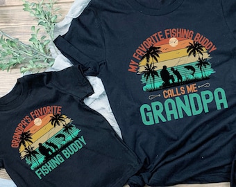May include: Two black t-shirts with a vintage sunset graphic and the text "Grandpa's Favorite Fishing Buddy" and "My Favorite Fishing Buddy Calls Me Grandpa".