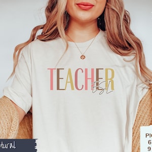 May include: A white t-shirt with the text "TEACHER ESL" in pink, brown, and yellow lettering. The text is in a modern font style. The shirt is being modeled by a person with long blonde hair.