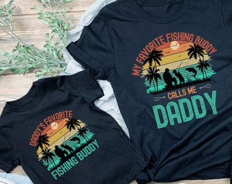 May include: Two black t-shirts with a retro sunset graphic and the text "Daddy's Favorite Fishing Buddy" and "My Favorite Fishing Buddy Calls Me Daddy".