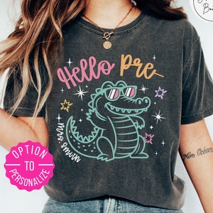 May include: A charcoal gray short-sleeve t-shirt featuring a graphic of a light green alligator wearing pink sunglasses. The text reads Hello Pre and Mrs. Smith in pink and orange script. A bright pink circular badge says Option to Personalize.