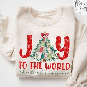 May include: Sand-colored sweatshirt with the word "JOY" in red, snowflake-decorated letters. A Christmas tree graphic is between the words. Below, the text reads "TO THE WORLD the Lord has come!"
