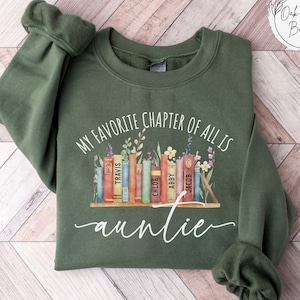 May include: Military green sweatshirt with the text "MY FAVORITE CHAPTER OF ALL IS auntie" in white script. The design features a row of colorful books with floral accents. The sweatshirt is a comfortable, casual style.