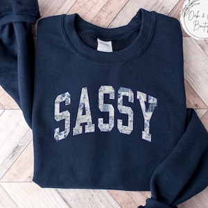 May include: Navy blue sweatshirt with the word "SASSY" in white, floral-patterned letters. The sweatshirt has a crew neck and long sleeves. The word "NAVY" is printed at the bottom left corner. The Oak & Olive Boutique logo is in the top right corner.