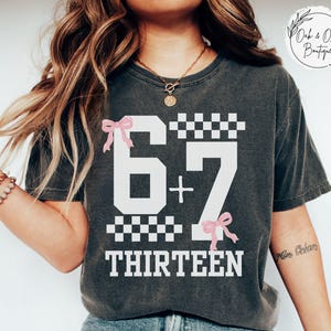 May include: Dark gray t-shirt with the numbers "67 + 7" and the word "THIRTEEN" in white. The design includes pink bows and checkered patterns. The word "PEPPER" is at the bottom.