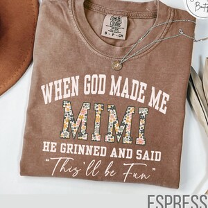 Puede incluir: Camiseta color espresso con el texto "WHEN GOD MADE ME MIMI HE GRINNED AND SAID 'This'll be Fun'" en blanco y estampado floral. La camiseta tiene cuello redondo y mangas cortas. Un collar plateado con un colgante est&aacute; drapeado sobre la camiseta.