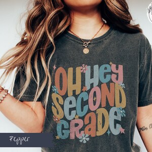 May include: A dark gray t-shirt with the text "Oh hey second grade" in colorful retro-style lettering. The shirt features a distressed look and is decorated with small flower graphics.
