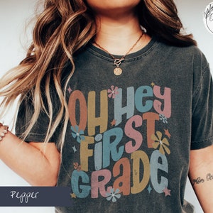 May include: A dark grey t-shirt with a colorful graphic that says "Oh Hey First Grade" with flowers and stars.