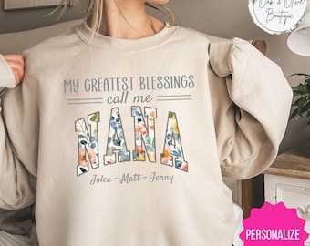 May include: A beige sweatshirt with a floral design. The text "My greatest blessings call me Nana" is printed on the front of the sweatshirt. The names "Jolee - Matt - Jenny" are printed below the text.