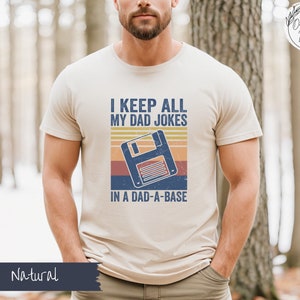 May include: A beige t-shirt with a retro-style graphic of a floppy disk and the text "I keep all my dad jokes in a dad-a-base".