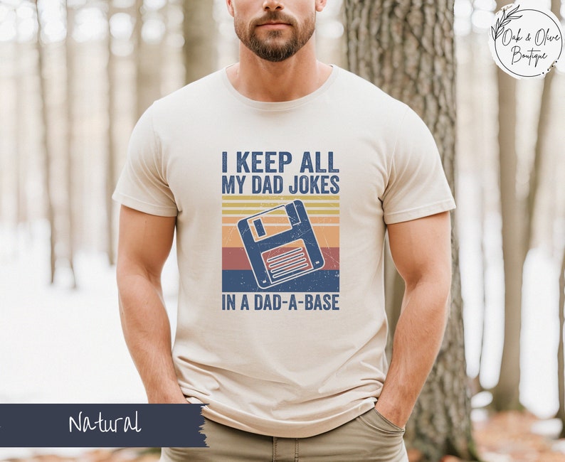 May include: A beige t-shirt with a retro-style graphic of a floppy disk and the text "I keep all my dad jokes in a dad-a-base".