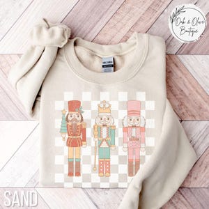 May include: A beige sweatshirt with a checkered pattern and three nutcracker illustrations. The nutcrackers are wearing red, green, and pink outfits. The text "SAND" is visible at the bottom of the image.