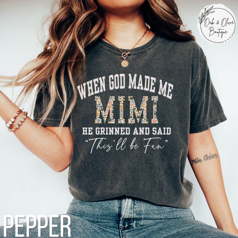 Puede incluir: Camiseta de color pimienta con el texto "WHEN GOD MADE ME MIMI HE GRINNED AND SAID 'This'll be Fun'". La palabra "MIMI" est&aacute; rellena con un estampado floral. La camiseta es de cuello redondo.
